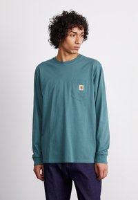 Camisa de manga larga en color turquesa con un bolsillo; fabricada en algodón; cuenta con un parche de logo cuadrado en el pecho; diseño ajustado con mangas con puños.