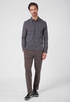 Langsleeve shirt in blauw met een geometrisch patroon van rechthoeken in bruin, beige en marineblauw; gecombineerd met bruin textuurbroek en donkere sneakers.