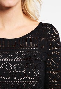 Femme portant un top en dentelle noire avec des motifs géométriques et floraux, featuring un large col rond et des manches longues.