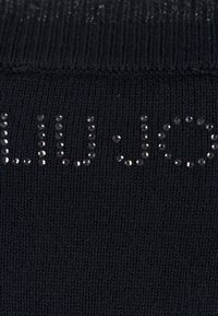 Tejido de punto negro con el nombre de la marca "LIU JO" hecho de pequeños brillantes negros dispuestos en una fuente estilizada.