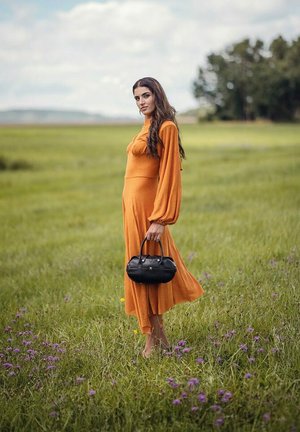 Oranžové šaty s dlouhým rukávem a upnutou sukňou, která volně splývá. Černá kabelka s zaobleným tvarem, nošená na travnatém poli s fialovými květinami.
