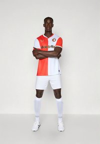 Rød og hvit fotballtrøye med delt design, rund hals, klubblogo og sponsor tekst. Brukes med hvite shorts og sokker.