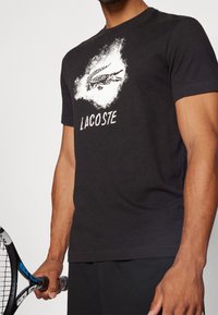 Mies, joka pitää yllä mustaa Lacoste T-paitaa, jossa on valkoinen krokotiililogo ja teksti, pitää mustaa ja sinistä tennismaila.