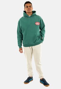 Sudadera de algodón verde con un bolsillo frontal y un logo rojo, combinada con pantalones de chándal beige claros y zapatillas azules con detalles.