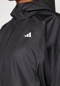 Gros plan d'une personne portant une veste noire zippée avec un petit logo blanc à trois bandes sur la poitrine.