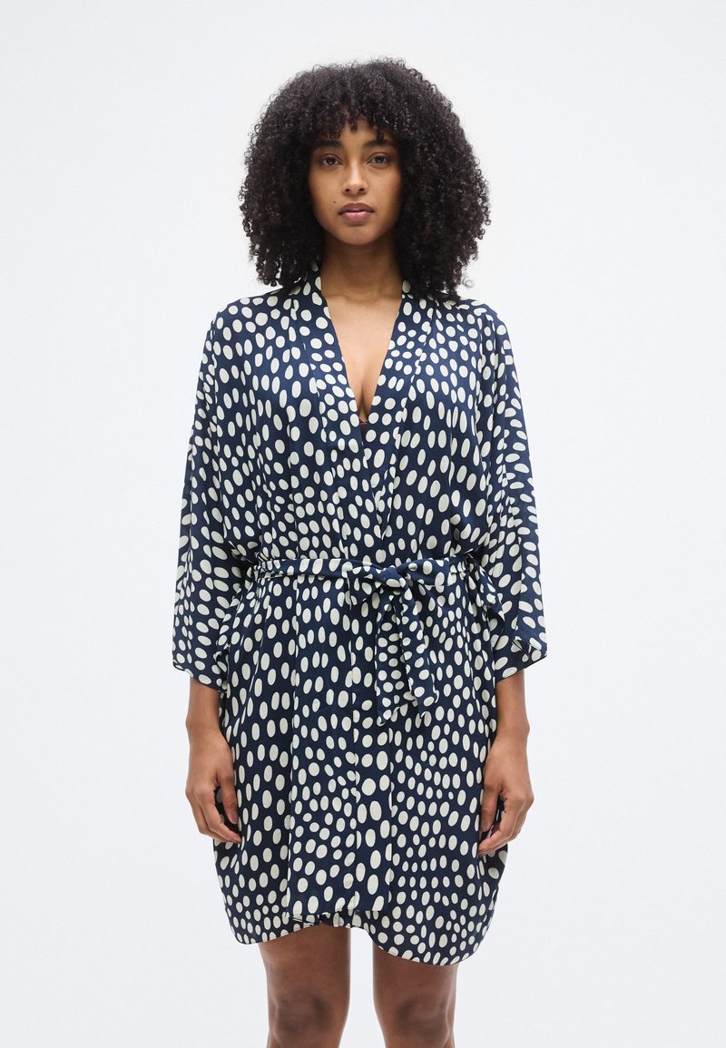 Μπλε ναυτικό μπουρνούζι με λευκό πουά σχέδιο, σχεδιασμένο με εμπρόσθιο τύλιγμα, φαρδιές μανσέτες και ζώνη στη μέση. Λείο, ελαφρύ ύφασμα.