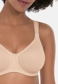 Beige bra with a textured pattern, V-shaped neckline, and decorative floral accent on the strap. Features padded cups and adjustable straps.