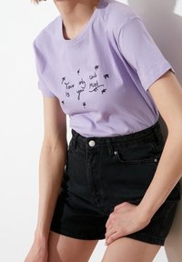 Camiseta de algodón lavanda con texto bordado en negro y pequeños gráficos florales. Combinada con pantalones cortos de mezclilla negra de tiro alto.