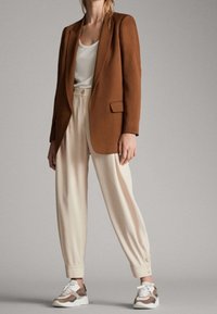 Brązowa marynarka, biały top, kremowe spodnie zwężane ku dołowi oraz sneakersy z brązowymi akcentami. Gładkie tekstury i dopasowany krój podkreślają nowoczesny strój.