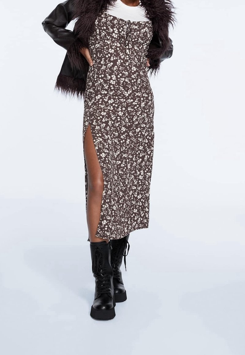 Femme portant une robe midi marron à motifs floraux avec une fente sur la cuisse, des bottes noires à lacets, un haut blanc et une veste en cuir noire à col en fourrure, mains sur les hanches.