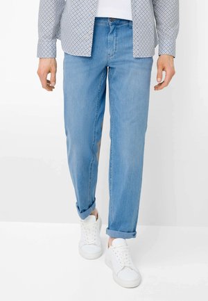 Mann trägt hellblaue hochgekrempelte Jeans, weiße Turnschuhe, ein gemustertes langärmeliges Hemd und ein weißes Unterhemd, steht vor weißem Hintergrund.