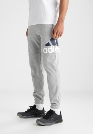 Graue Jogginghosen mit elastischen Bündchen, ausgestattet mit einem großen weißen Adidas-Logo und marineblauen Akzenten, kombiniert mit schwarzen Sportschuhen.