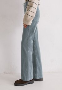 Lichtblauwe wijde broek van zachte stof, met voorzakken en een nette afwerking. Draagt met donkerbruine schoenen.
