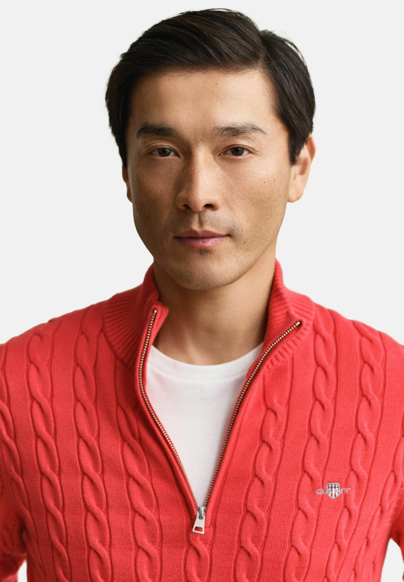 Uomo con capelli corti e scuri che indossa un maglione rosso a trecce con cerniera sopra una camicia bianca, rivolto verso la fotocamera con un'espressione neutra.