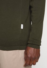 Olivgrüner Strickpullover mit einer strukturierten Bündchen am Saum und einem kleinen Markentag an der Seite. Sichtbare Hand, die den Stoff greift.