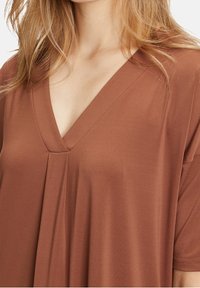 Brun V-ringad topp gjord av mjukt tyg, med lös passform och subtil plissering vid halsringningen, korta ärmar och slät yta.