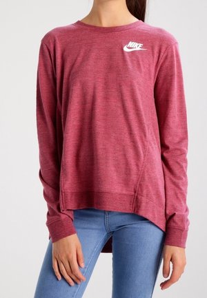 Femme portant un sweat-shirt Nike à manches longues couleur bordeaux, associé à un jean bleu clair, debout les mains détendues le long du corps.