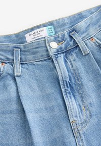 Jeans palazzo de tiro alto en azul claro y de pierna ancha. Presenta costuras superiores, un botón frontal y una etiqueta que muestra los detalles del tamaño. Textura de denim visible.