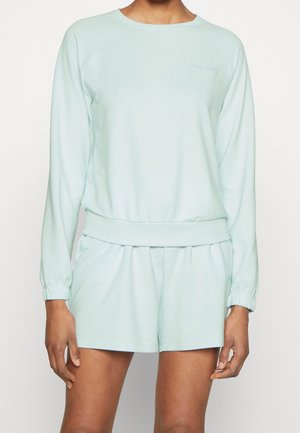 Person iført lyseblå langærmet sweatshirt og matchende shorts med elastiske manchetter og taljebånd, stående mod en ensfarvet baggrund.