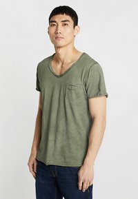 T-shirt in cotone verde oliva con scollo a V e maniche arrotolate, dotata di una piccola tasca sul petto e una vestibilità comoda. Il tessuto ha una finitura leggermente texture.