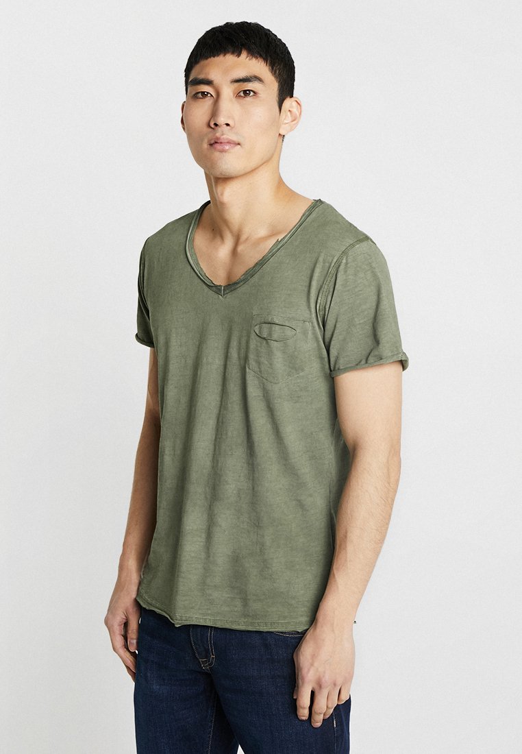 T-shirt in cotone verde oliva con scollo a V e maniche arrotolate, dotata di una piccola tasca sul petto e una vestibilità comoda. Il tessuto ha una finitura leggermente texture.