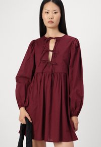 Vestido borgonha com mangas bufantes longas, decote em forma de fenda com atilhos e saia rodada, feito de tecido leve.