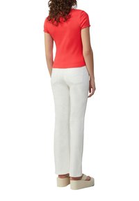 Vrouw in een aansluitende rode top met korte mouwen, witte rechte broek en beige plateau-sandalen, staand met haar rug naar de kijker.
