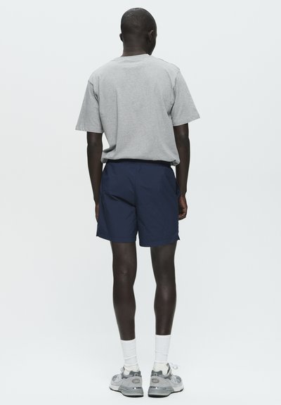 T-shirt gris à manches courtes associé à un short marine, présenté de dos. Le modèle porte des baskets grises et des chaussettes blanches, sur fond épuré.