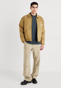 Giacca in tela marrone con una tasca, t-shirt grigia, pantaloni cargo beige e stivali neri con cuciture gialle, su uno sfondo neutro.