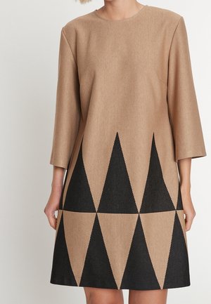 Woman wearing a tan dress with long sleeves and large black geometric triangle patterns along the bottom half.