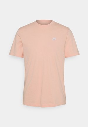 T-shirt en coton pêche clair avec un col rond, des manches courtes et un logo Nike blanc sur le côté gauche de la poitrine. Texture lisse, coupe décontractée.