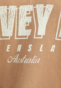 T-shirt en tissu brun avec un grand texte blanc usé. "HARVEY" est en évidence, avec "ENSLA" et "Australia" en texte cursif plus petit en dessous.