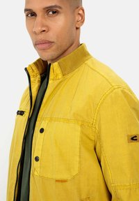 Gelbe Jacke mit strukturierter Oberfläche, Reißverschluss, geripptem Kragen und Brusttaschen. Besitzt Kontrastnähte und ein Logopatch am Ärmel.