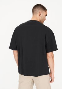 Camiseta negra de manga corta con un corte relajado, escote redondo y textura suave, mostrada desde la parte de atrás. Se llevan pantalones de color claro.