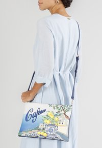 Vit crossbody-väska med ett färgglatt citronmotiv, blå accenter och tryckt text. Tillverkad av ett mjukt material, med en avtagbar rem.