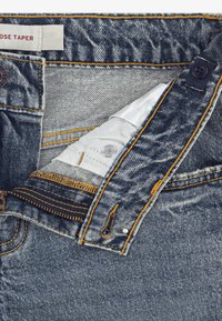 Τζιν τζιν με κωνική εφαρμογή, με αντίθετες πορτοκαλί ραφές, κλείσιμο με κουμπί και μεταλλική λεπτομέρεια τσέπης για κέρματα.