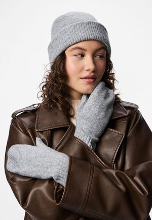 Kvinna i brun läderjacka, ljusgrå stickad mössa och matchande vantar, poserar med en hand nära ansiktet mot en enfärgad bakgrund.