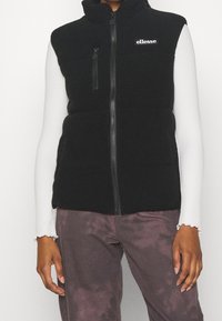Persona che indossa un gilet in fleece nero con il logo "ellesse", una maglietta a maniche lunghe bianca e pantaloni viola sbiaditi, in piedi contro uno sfondo bianco.