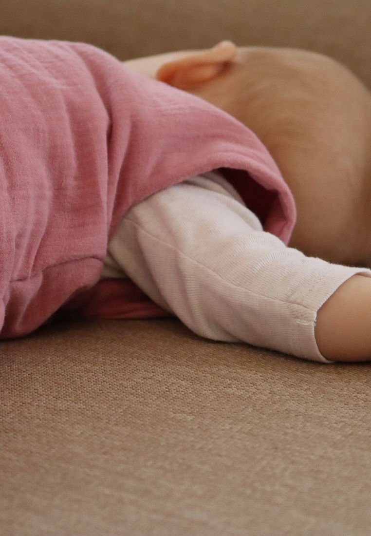 Ein schlafendes Baby, das ein rosa Kleidungsstück über einem hellen Langarmshirt trägt, ruht auf einer strukturierten beigen Oberfläche.