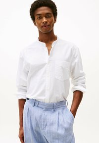 Camicia bianca a maniche lunghe con colletto alla cinese, caratterizzata da un motivo testurizzato e una tasca sul petto, abbinata a pantaloni plissettati blu chiaro.