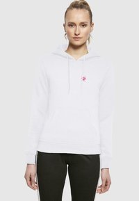 Felpa bianca con logo rosa, cappuccio con cordino, tasca anteriore a marsupio e polsini a coste. Abbinata a pantaloni neri con strisce bianche.