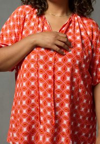 Red and white patterned blouse with a V-neck, gathered neckline, short sleeves, and a loose fit. Features a geometric design with curved shapes.