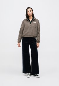 Brązowa bluza polarowa z półzamknięciem i logo Gap, zestawiona z czarnymi szerokimi spodniami sztruksowymi oraz czarno-białymi trampkami.