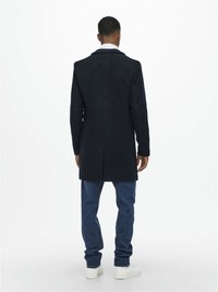 Manteau en laine mélangée navy avec une coupe ajustée, une fente au dos, et un revers cranté, associé à un jean en denim noir et des baskets blanches.
