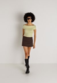 Top off-shoulder de rayas en verde claro combinado con una mini falda a rayas marrón. La modelo lleva botas altas negras y gafas de sol, de pie contra un fondo liso.
