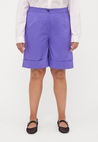 Pantaloni corti viola con orli piegati, realizzati in tessuto morbido. Presentano tasche laterali e una chiusura con bottone, abbinati a scarpe in rete nere.