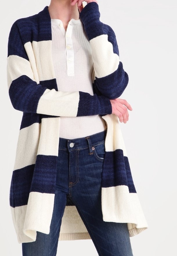 En kvinna som bär en marinblå och krämfärgad randig kardiagan över en ljus topp, med handen nära hakan, står mot en enkel bakgrund.