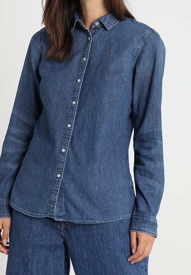 Jeansskjorta i mörkblått med krage, långa ärmar, knappar fram och en liten logotyp på bröstet. Slät textur.