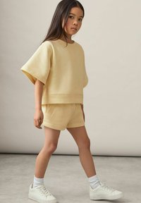 Gul bomuldsblandings sweatshirt med brede ærmer, parret med matchende shorts. Hvite sneakers og knæhøje sokker fuldender outfittet.
