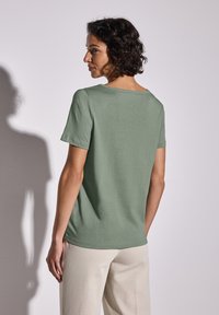 Světle zelené bavlněné tričko s uvolněným střihem, krátkými rukávy a širokým výstřihem, zobrazené ze zadní strany, v kombinaci s béžovými kalhotami.